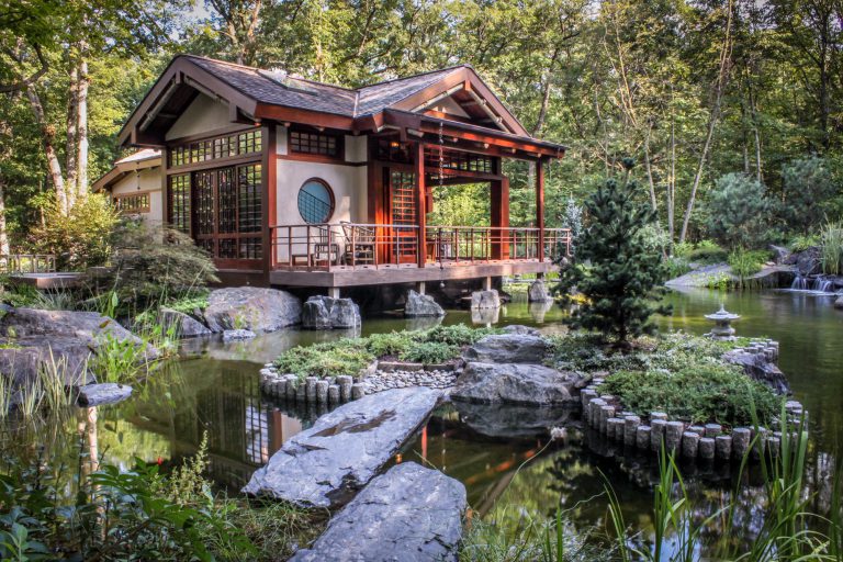 A weathered stone bridge arches over a tranquil koi pond, its still waters reflecting the warmth of a handcrafted Japanese tea house nestled among ancient garden plantings — Kane Brothers luxury Asian-inspired landscape design